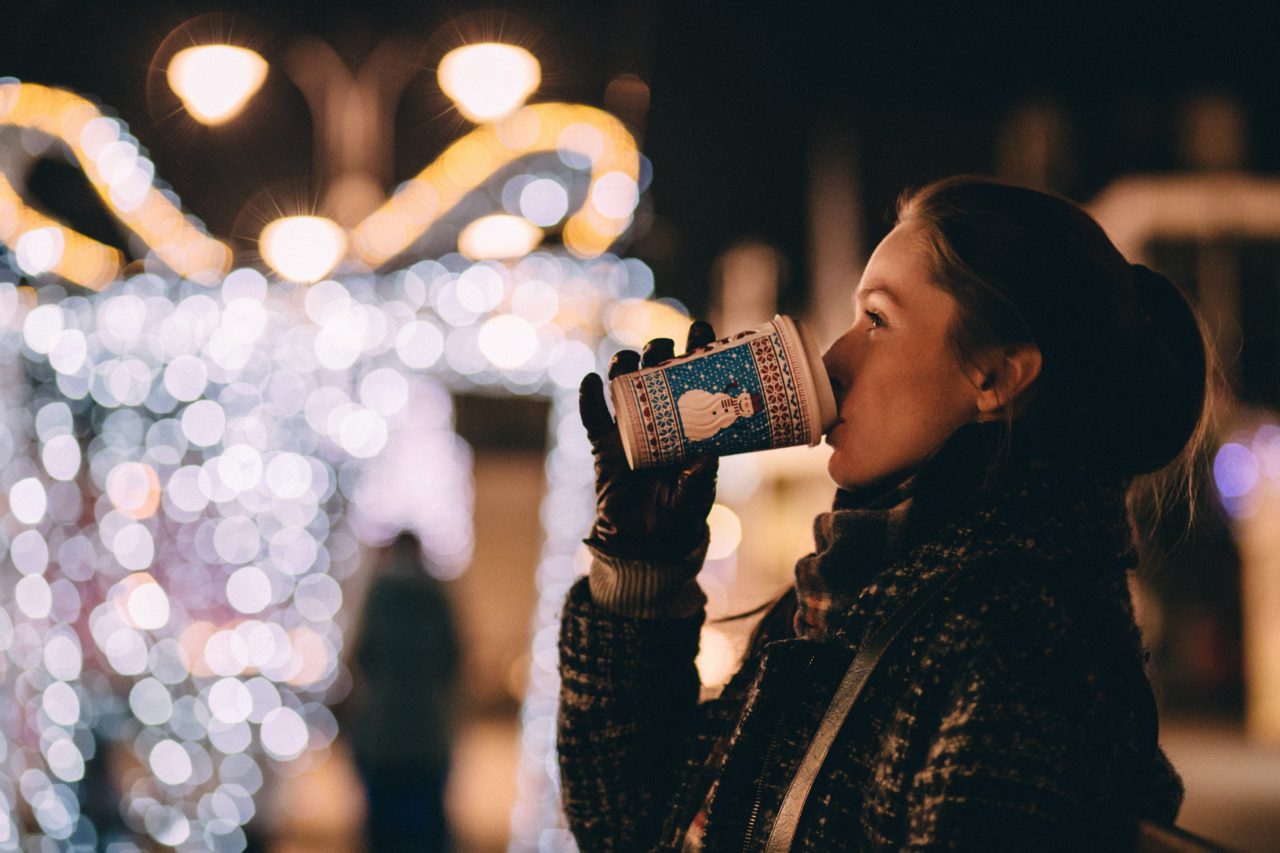 Jeune femme joyeuse en gants dégustant une boisson ornée d'un bonhomme de neige, tandis qu'au second plan, un bokeh lumineux aux couleurs de Noël crée une ambiance chaleureuse au cœur d'un marché de Noël.